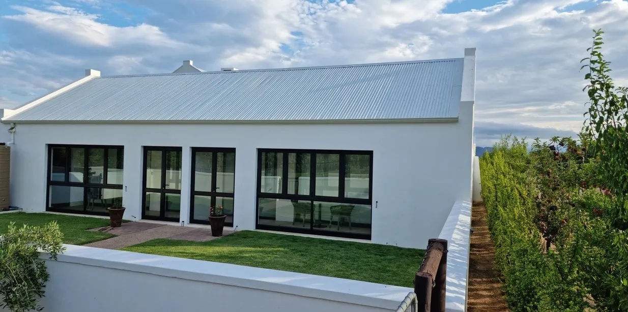Exterior of the Blue Strings Room at Klawerjas, whitewashed Cape Dutch-style building with dark-framed doors opening onto a lawned garden, vineyard rows alongside